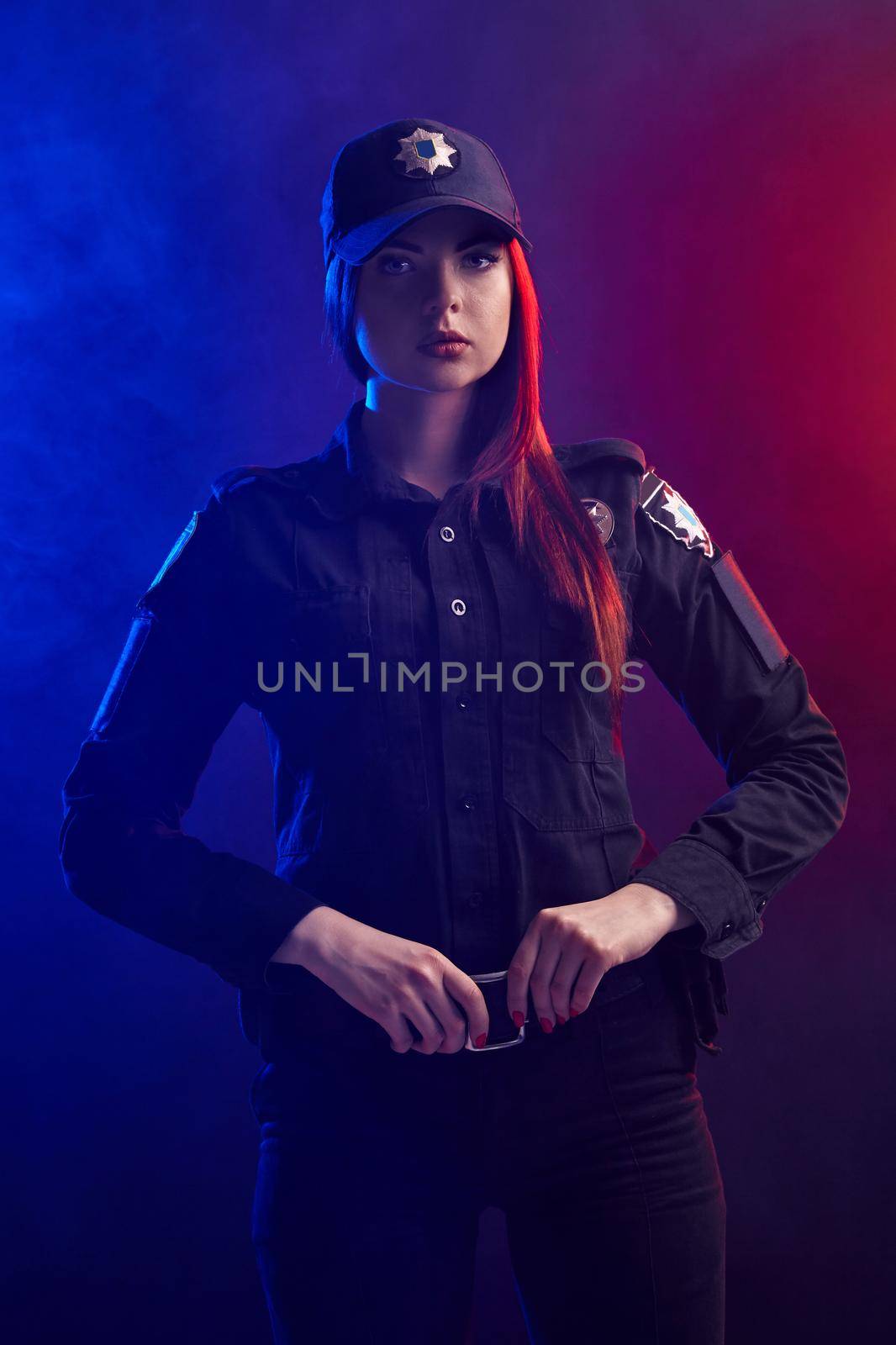 Serious redheaded female police officer in a uniform and a cap, with bright make-up, is holding her belt and looking at the camera against a black background with red and blue backlighting.