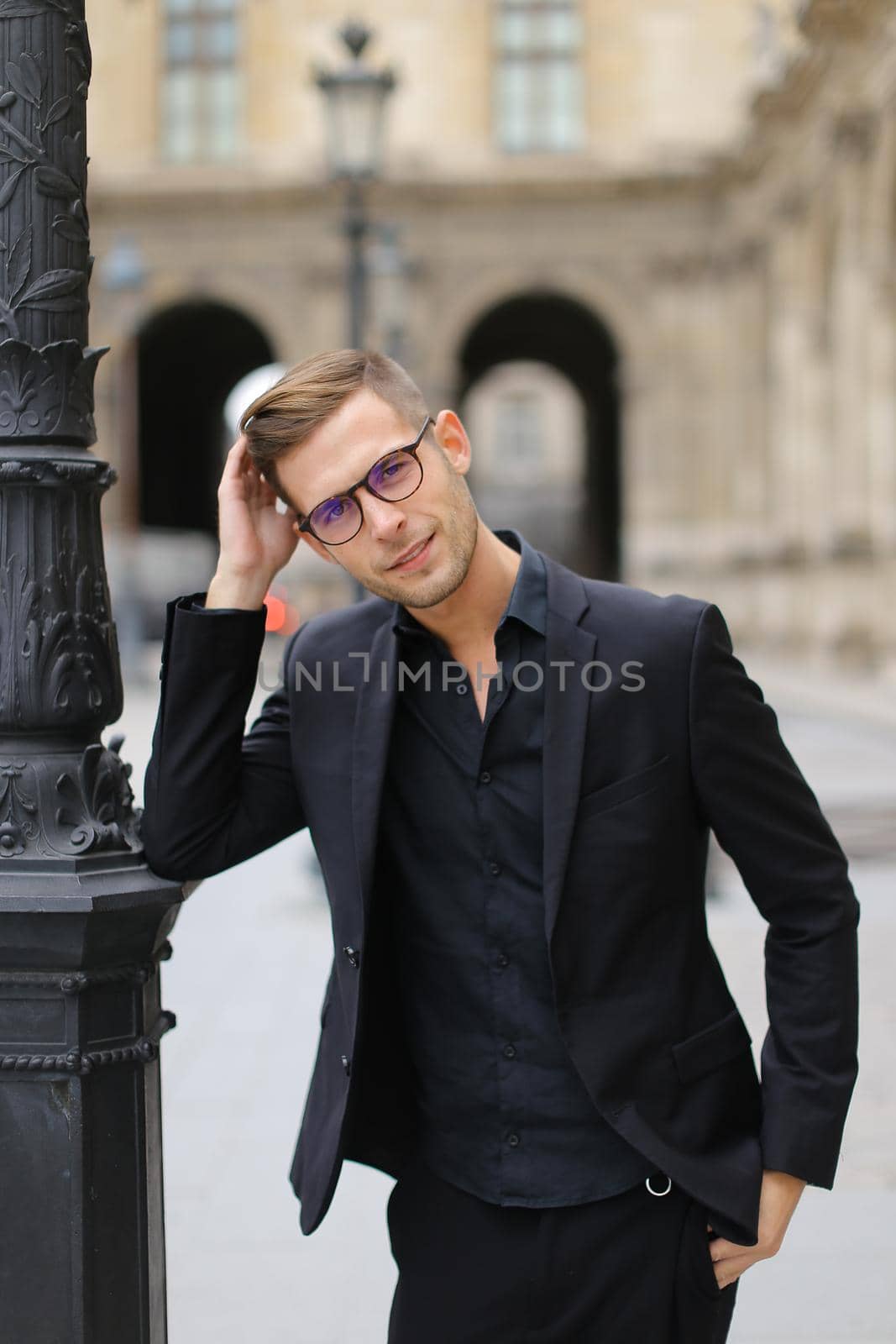 Young man in glasses walking in city and wearing black suit, leaning on lantern. Concept of strolling and male fashion model, urban photo session.