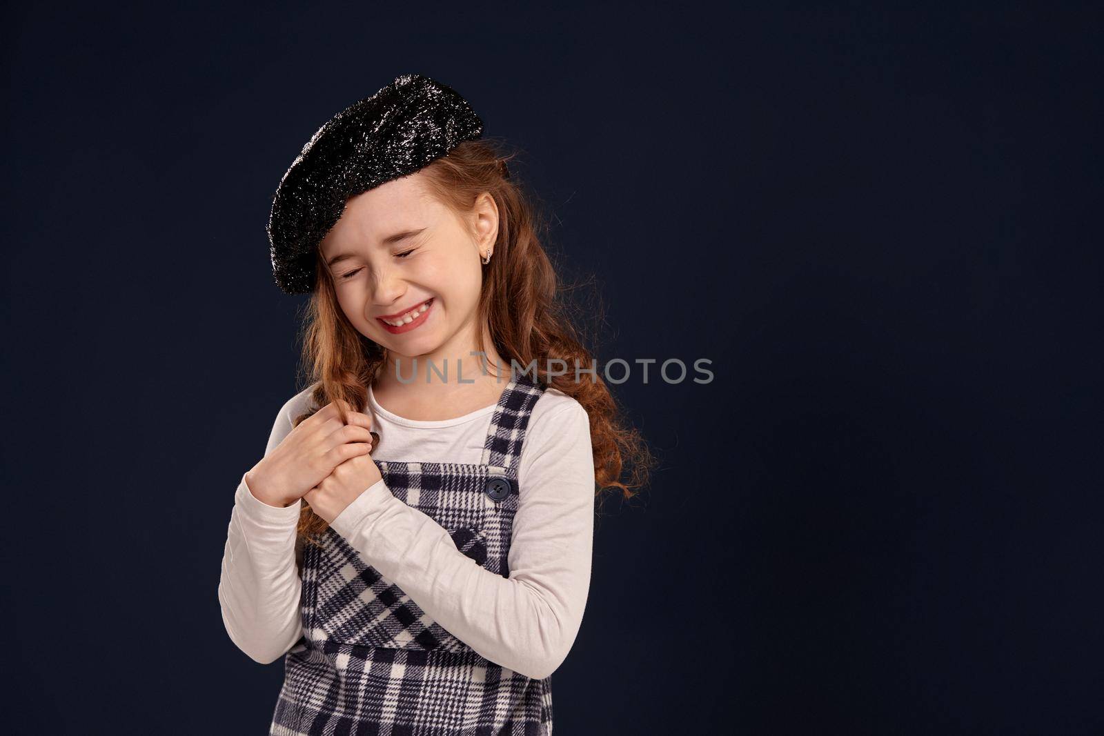Emotional little girl with a beautiful curly hair wearing checkered dress, white blouse and a black sparkling beret has closed her eyes and laughing. Stylish brunette kid is posing in studio on a black background. Children's fashion.