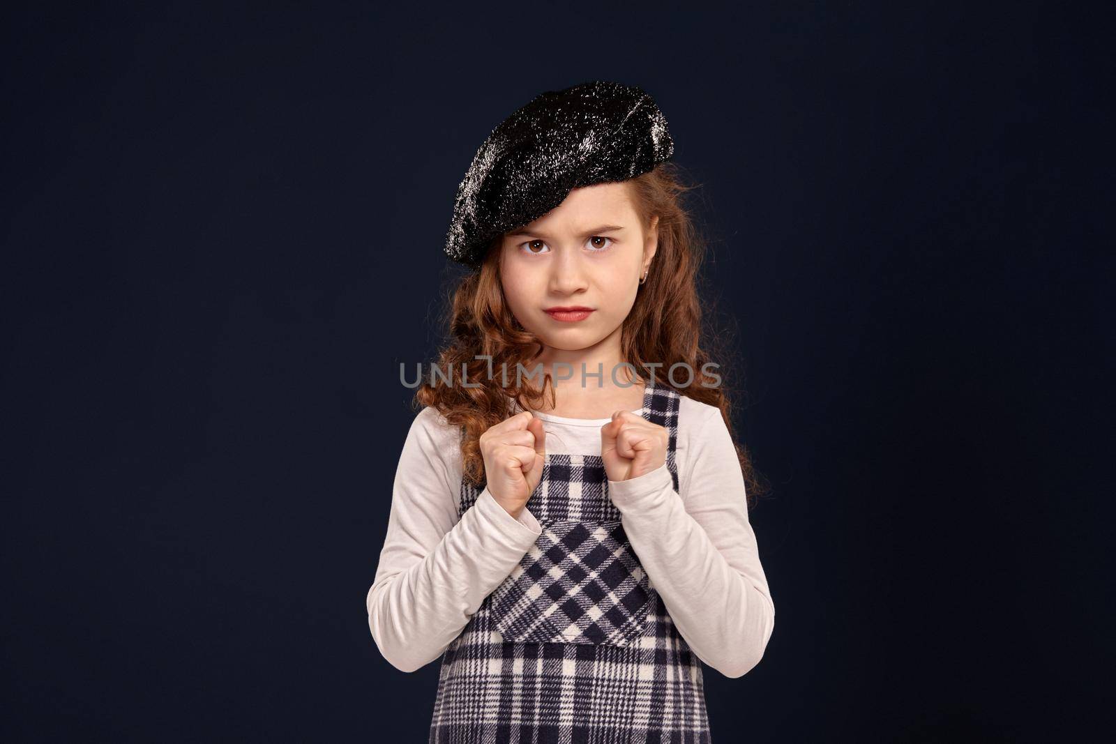 Unsmile little girl with a beautiful curly hair wearing checkered dress, white blouse and a black sparkling beret has clenched her fists and looking at the camera. Stylish brunette kid is posing in studio on a black background. Children's fashion.