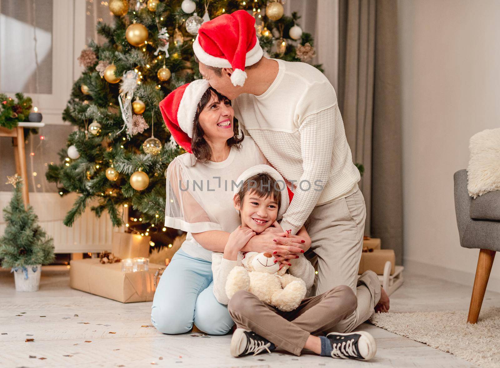 Happy family in santa hats sitting on floor under decorated christmas tree at home