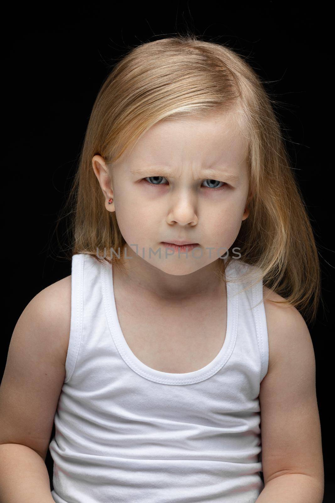 Serious toddler girl frowning and looking at the camera while sitting on black background in white undershirt. Childhood concept