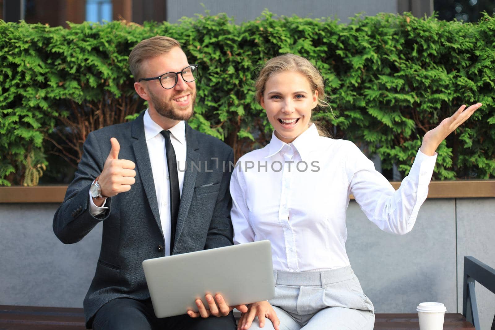 Office colleagues using laptop computer and showing thumbs up while sitting on a bench outdoor
