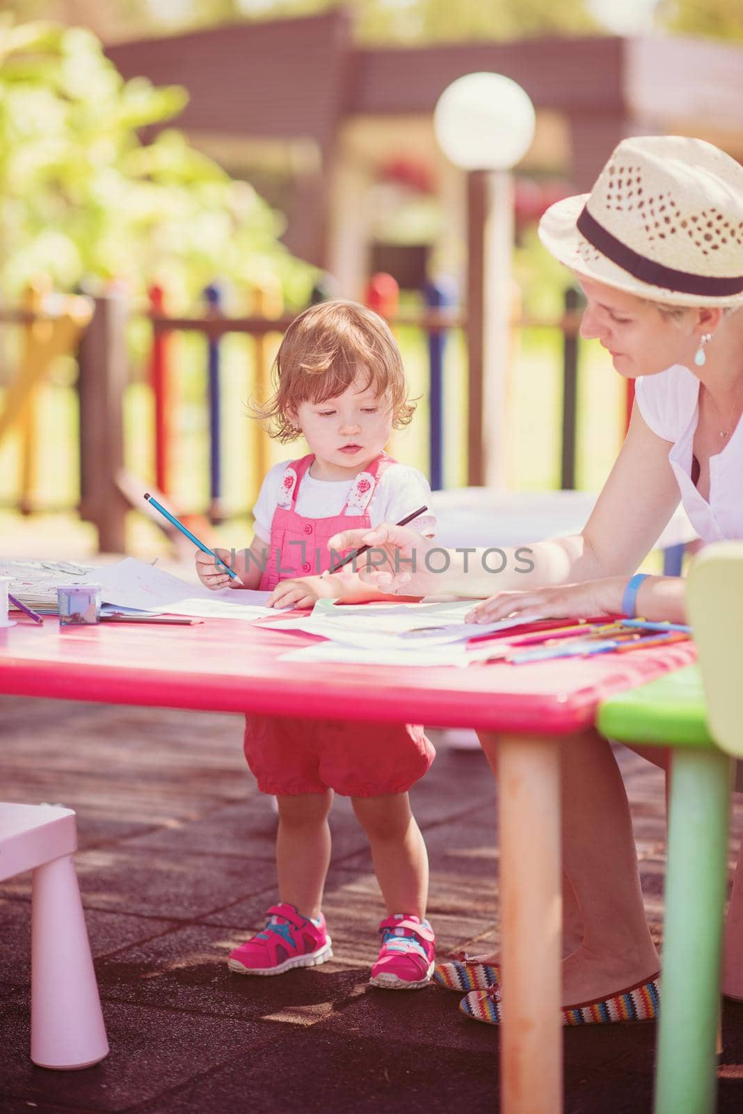 young mother and her little daughter cheerfully spending time together using pencil crayons while drawing a colorful pictures in the outside playschool