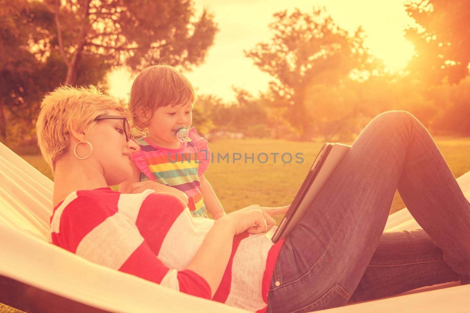 Happy mother and her little daughter enjoying free time using tablet computer while relaxing in a hammock during sunny day on holiday home garden