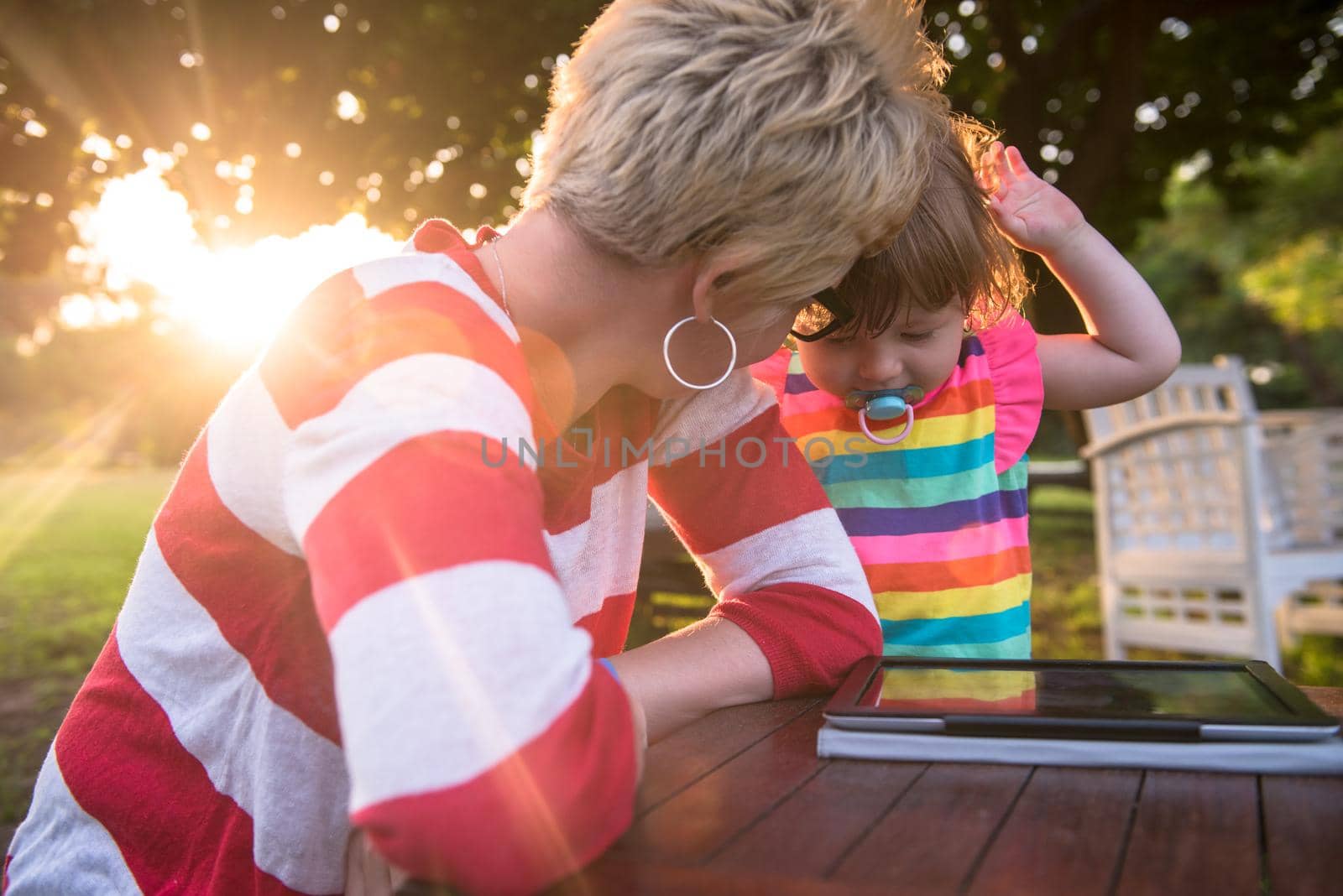 Happy mother and her little daughter enjoying free time using tablet computer while relaxing  on holiday home garden during sunny day