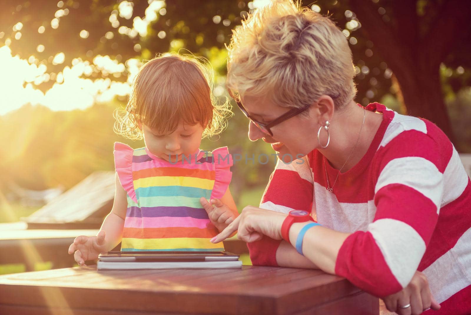 Happy mother and her little daughter enjoying free time using tablet computer while relaxing  on holiday home garden during sunny day