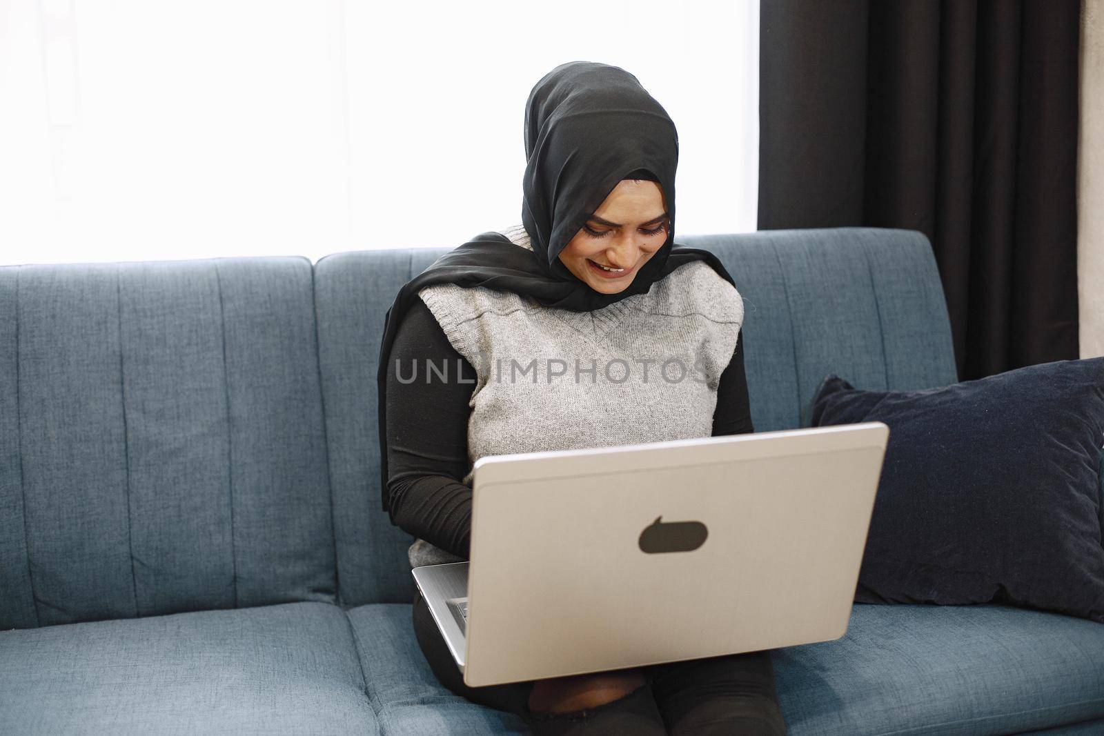 Modern arab girl in hijab using laptop at home, working remotely while sitting on couch in living room