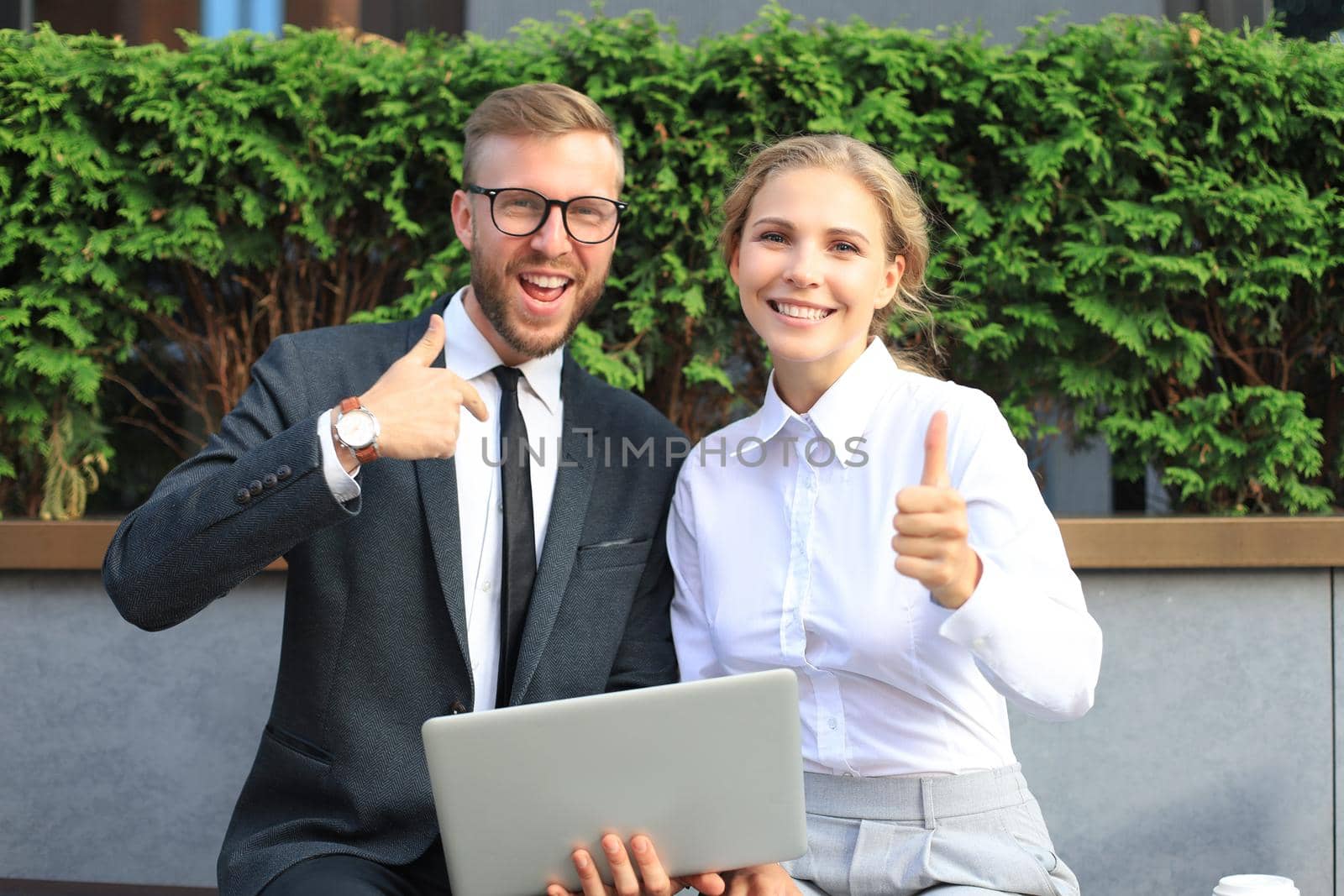 Office colleagues using laptop computer and showing thumbs up while sitting on a bench outdoor