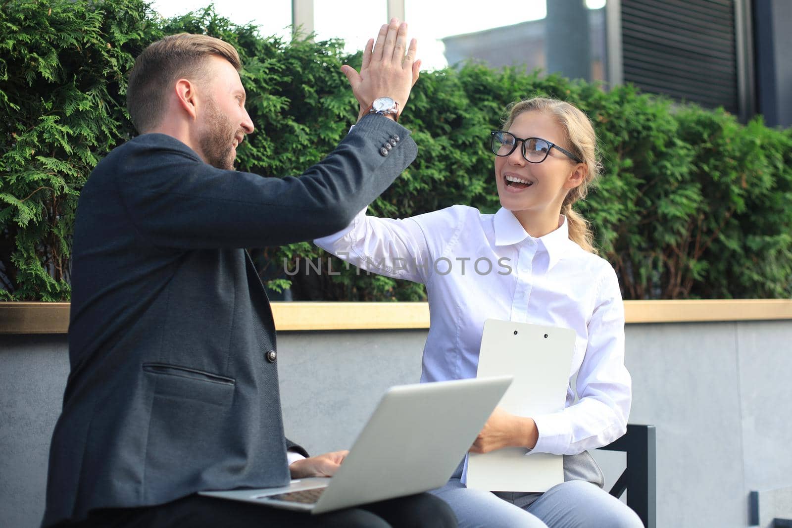 Office colleagues using laptop computer and giving five while sitting on a bench outdoor