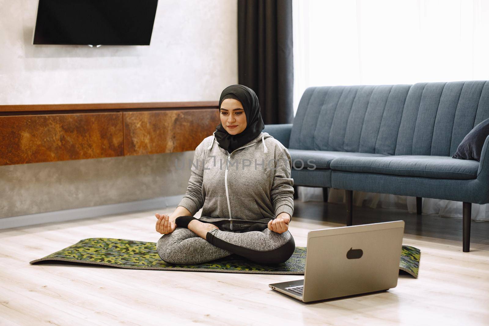 Home sport for muslim women. Smiling arabic girl in hijab practicing online yoga, watching video tutorial on laptop, excersising in living room