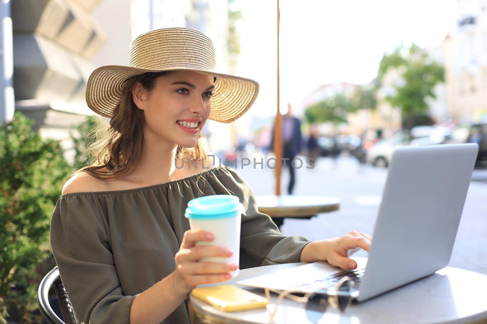 Happy nice woman working on laptop in street cafe, holding paper cup