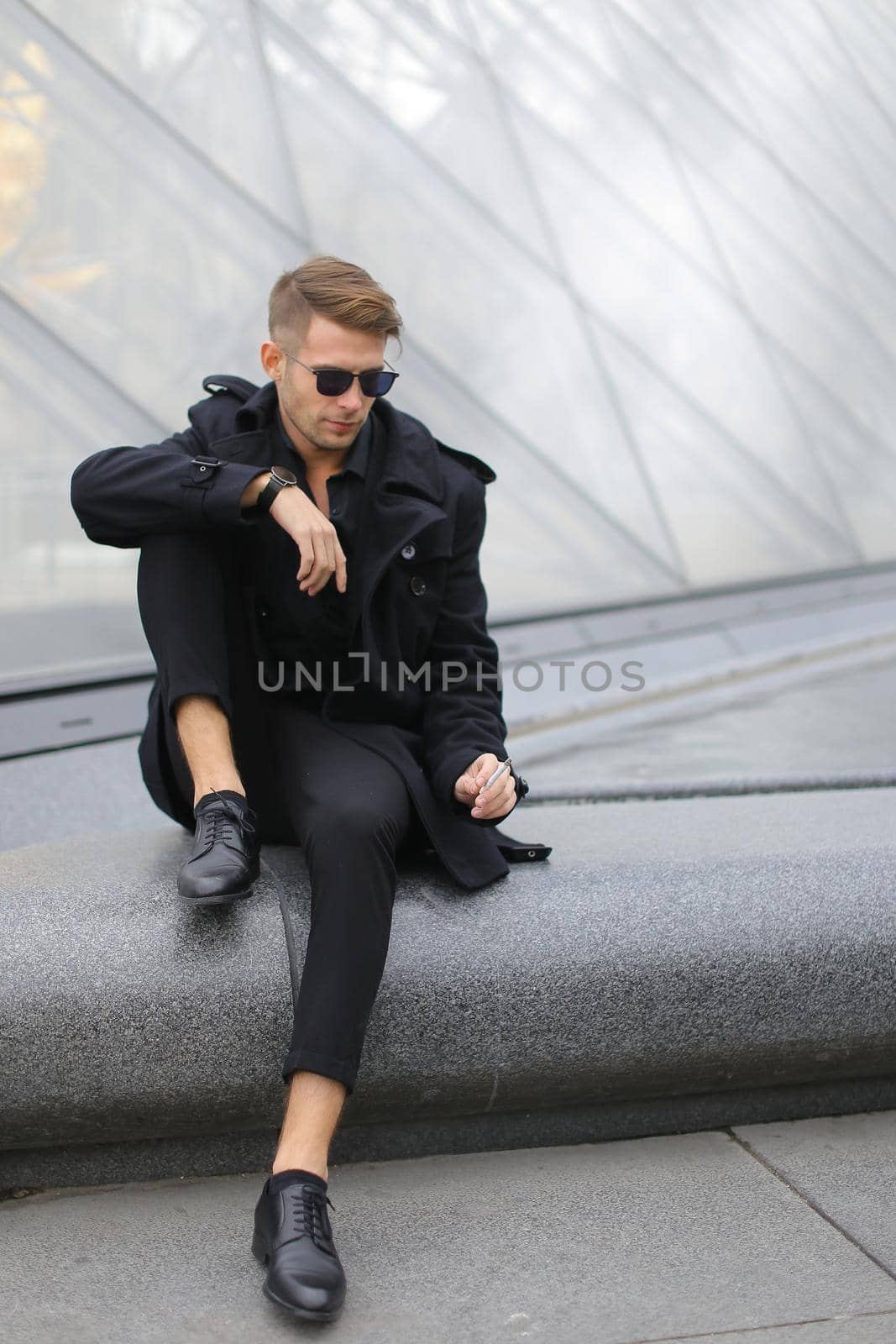 Young european man wearing black jacket sitting on Louvre Pyramid and smoking. by sisterspro