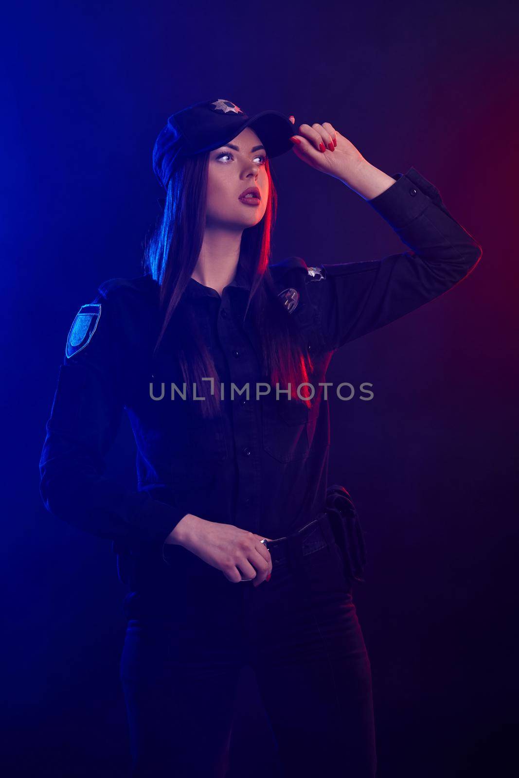 Redheaded lady police officer in a dark uniform, with bright make-up, is holding her cap looking away against a black background with red and blue backlighting. Defender of citizens is ready to enforce a law and stop a crime.