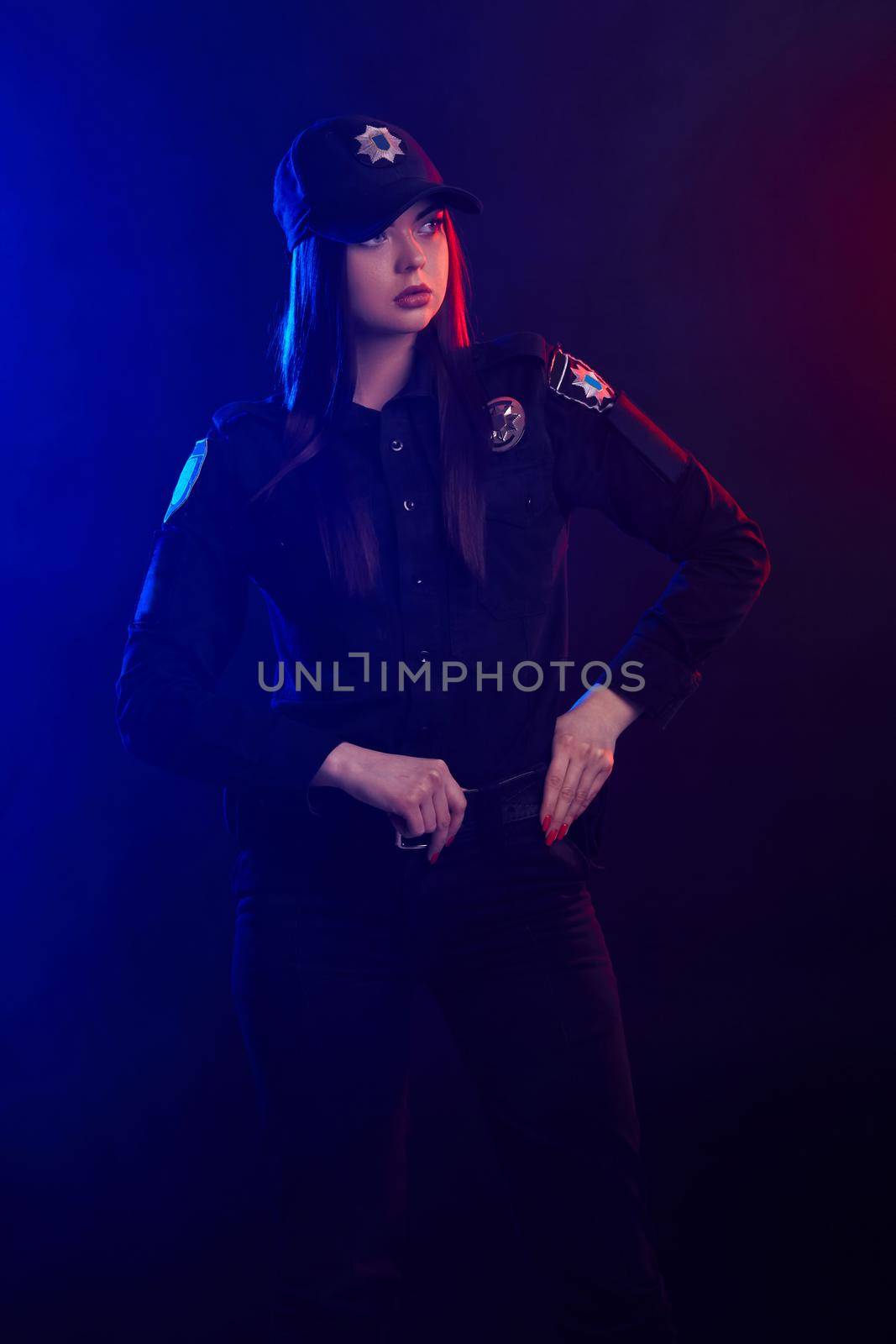 Serious female police officer is posing for the camera against a black background with red and blue backlighting. by nazarovsergey