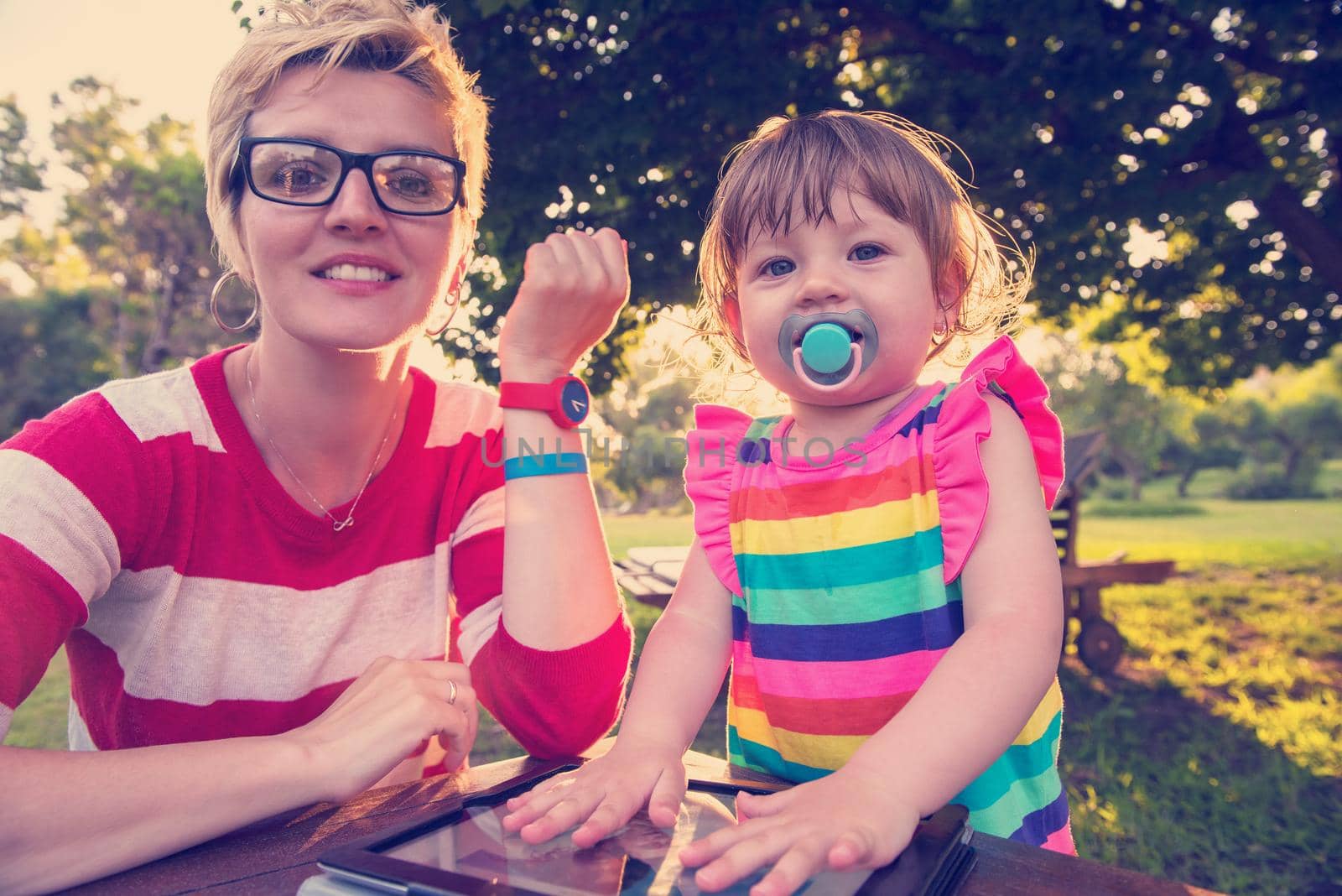 Happy mother and her little daughter enjoying free time using tablet computer while relaxing  on holiday home garden during sunny day