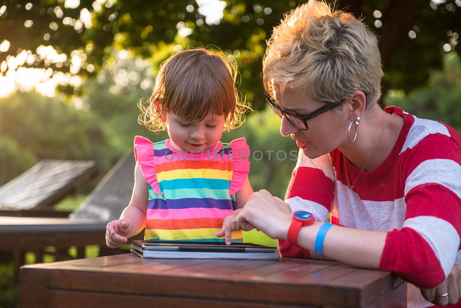 Happy mother and her little daughter enjoying free time using tablet computer while relaxing  on holiday home garden during sunny day