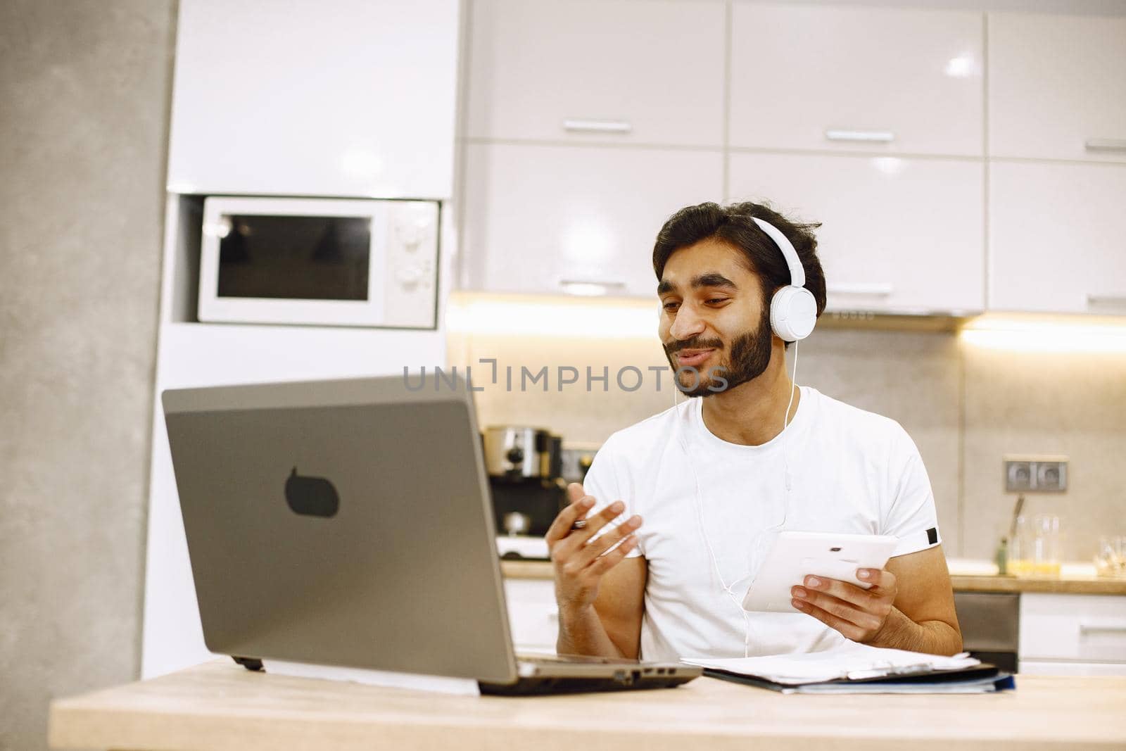 Arabic man watching online webinar, sitting in a kitchen with computer, enjoying distance learning.