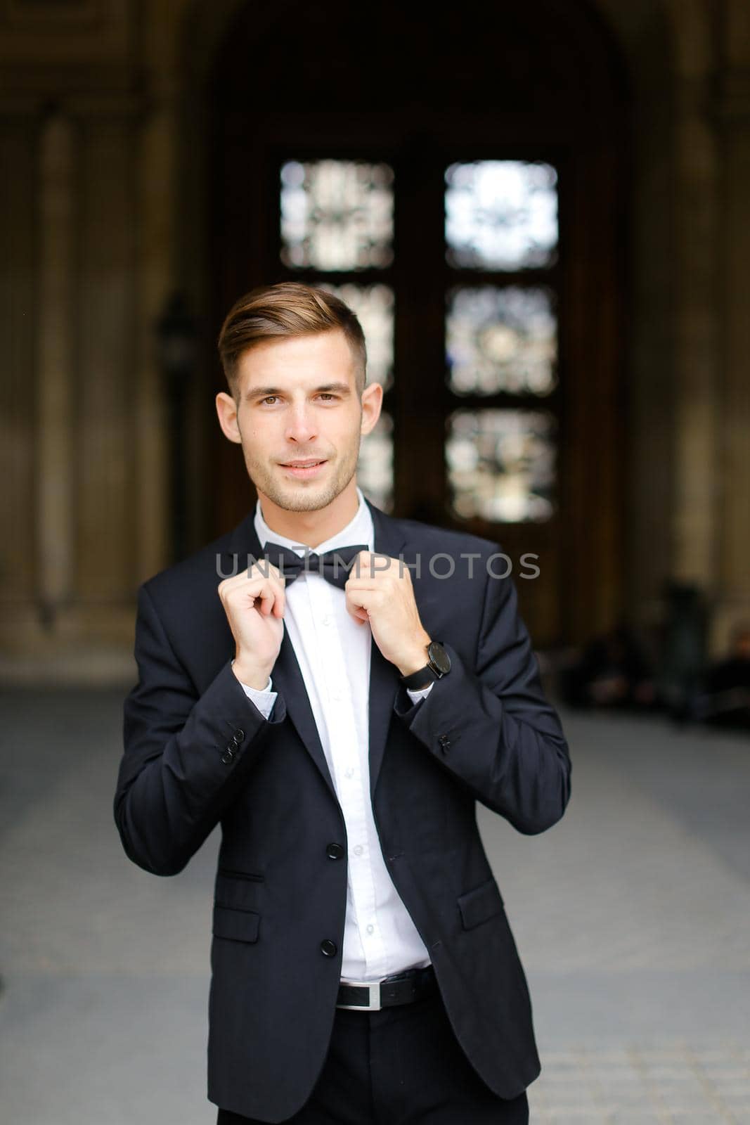 Young caucaisian man standing and posing, wearing black suit and bow tie, window in background. Concept of fashionable businessman and male model.