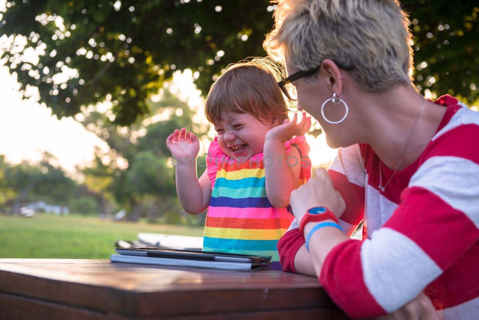 Happy mother and her little daughter enjoying free time using tablet computer while relaxing  on holiday home garden during sunny day