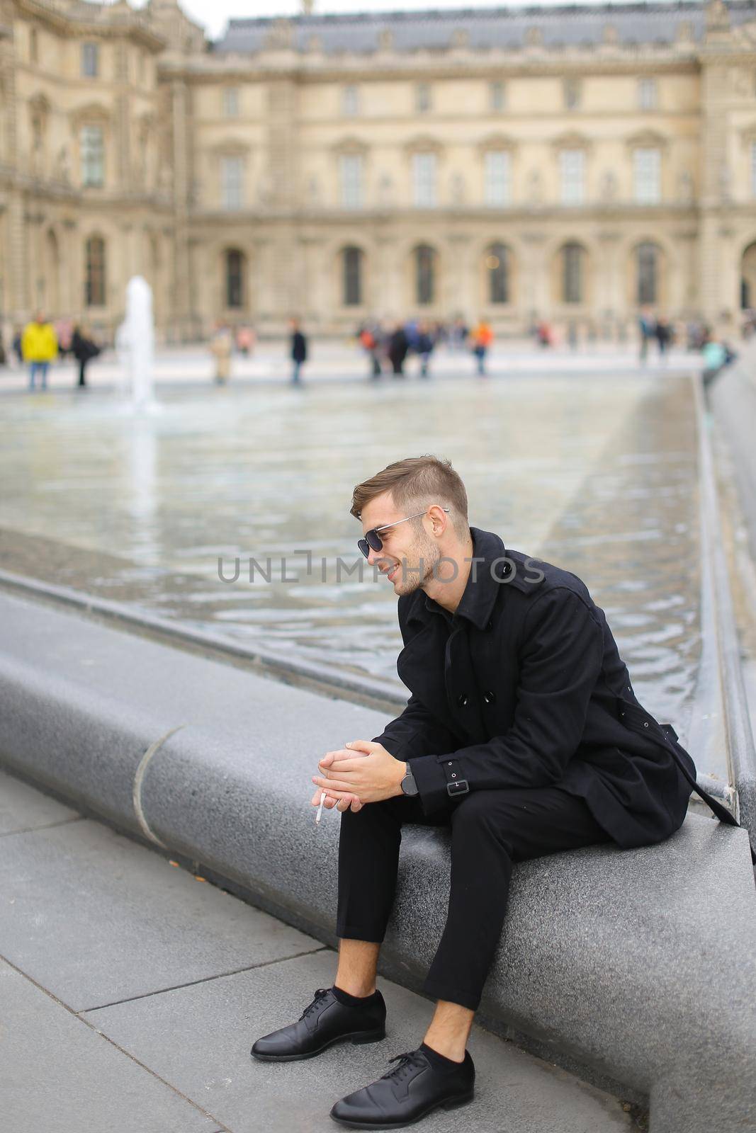Young man wearing black suit sitting near fountain and smoking cigarette in Paris, France. Concept of urban youth and male model for photo session in open air.