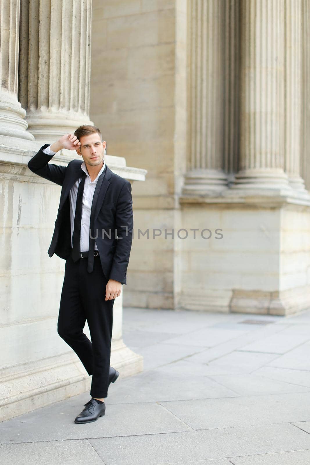 Young handsome man wearing black suit and tie leaning on column of building. by sisterspro