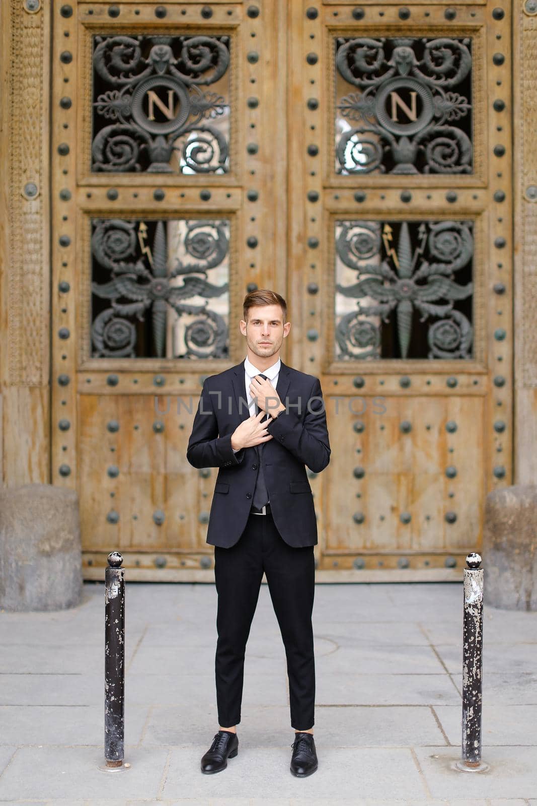 Handsome cauasian groom standing near wooden door and straightening tie. by sisterspro