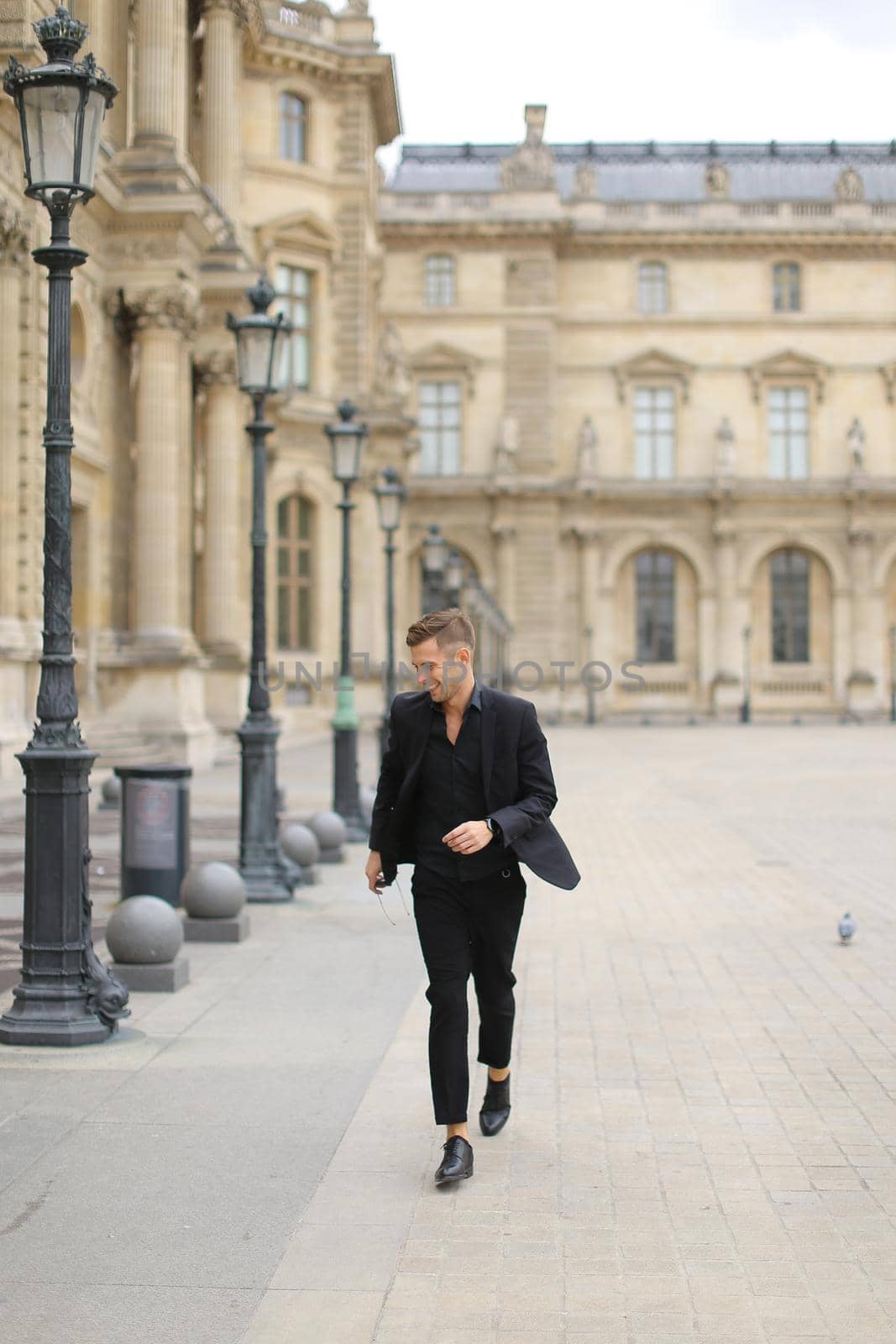 Young man strolling near building in Parisand wearing black suit, lanterns in background. Concept of male fashion model and urban photo session in France.