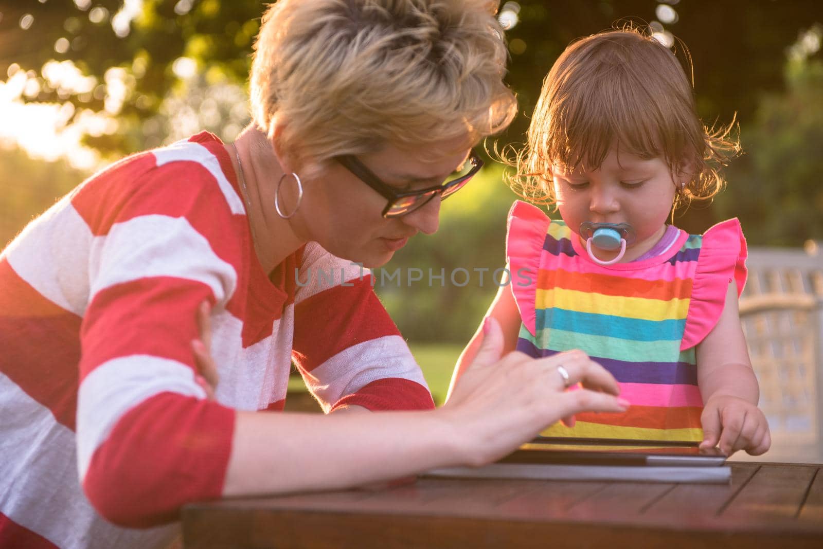 Happy mother and her little daughter enjoying free time using tablet computer while relaxing  on holiday home garden during sunny day