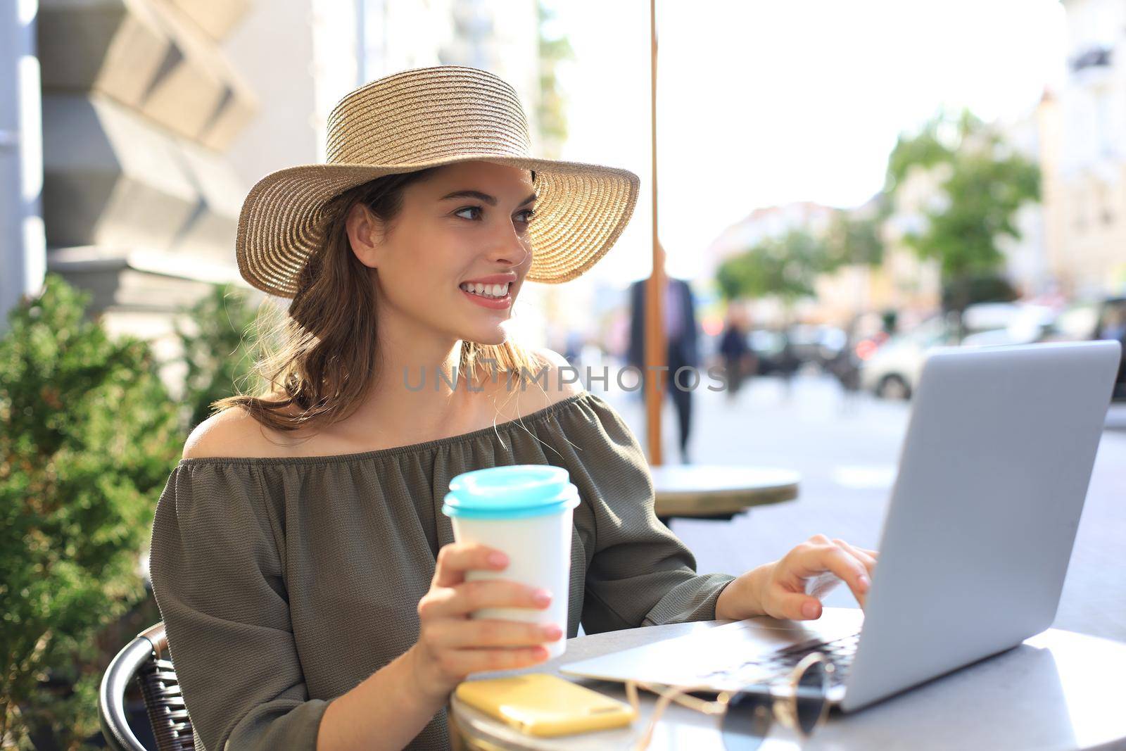 Happy nice woman working on laptop in street cafe, holding paper cup