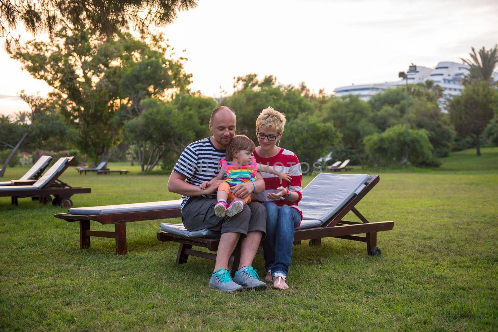 portrait of young happy family with a little daughter  sitting on the sunbed in the beautiful tropical park