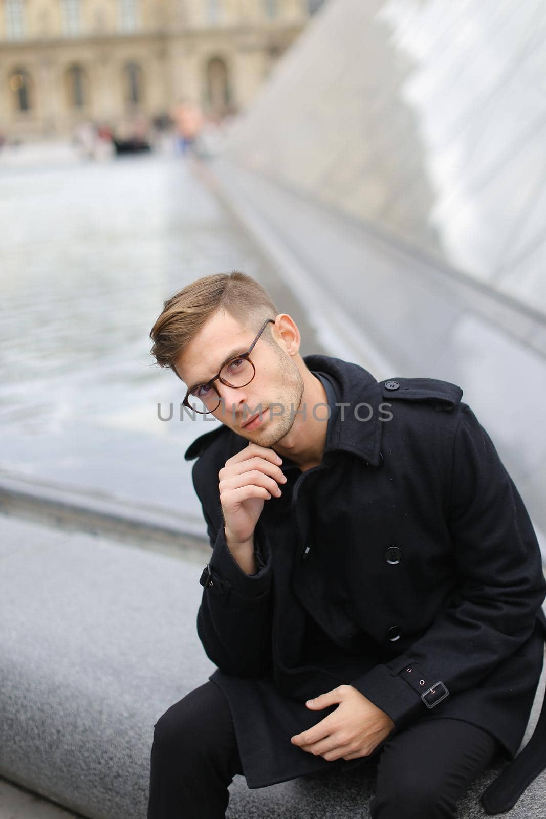 Young man wearing black jacket sitting on glass Louvre Pyramid. by sisterspro