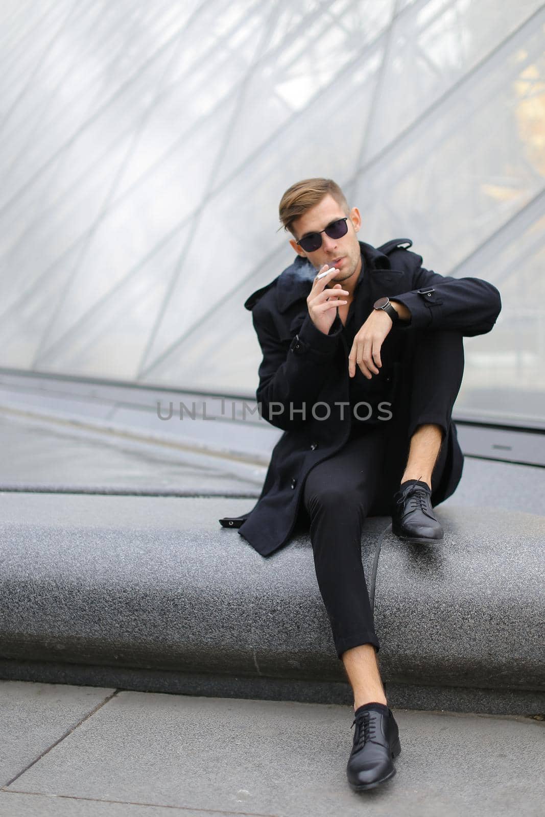 Young blond man in sunglasses wearing black jacket sitting near glass Louvre Pyramid in Paris and smoking cigarette, France. Concept of male fashion model and urban photo session.