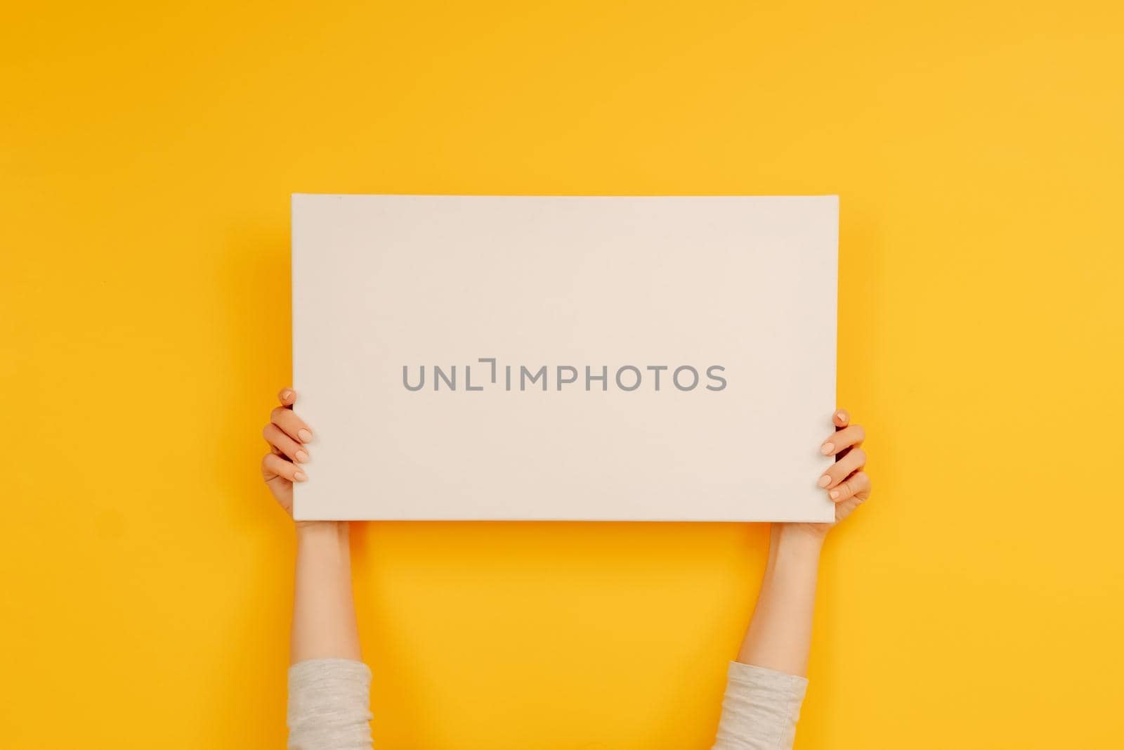 Hands holding white canvas paper blank with copyspace on yellow background. Empty sheet banner with place for text. Concept of design and creativity