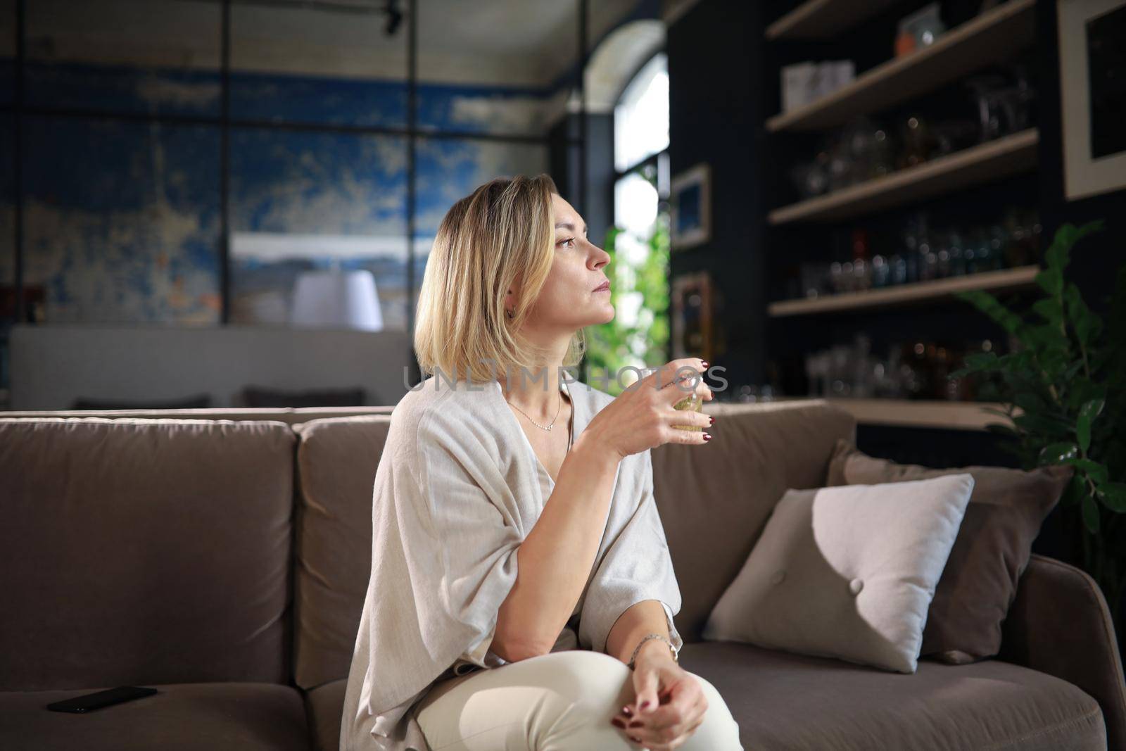 Happy mature woman resting on comfortable sofa drink coffee or tea, looking away, relaxing on cozy couch at home enjoy hot beverage