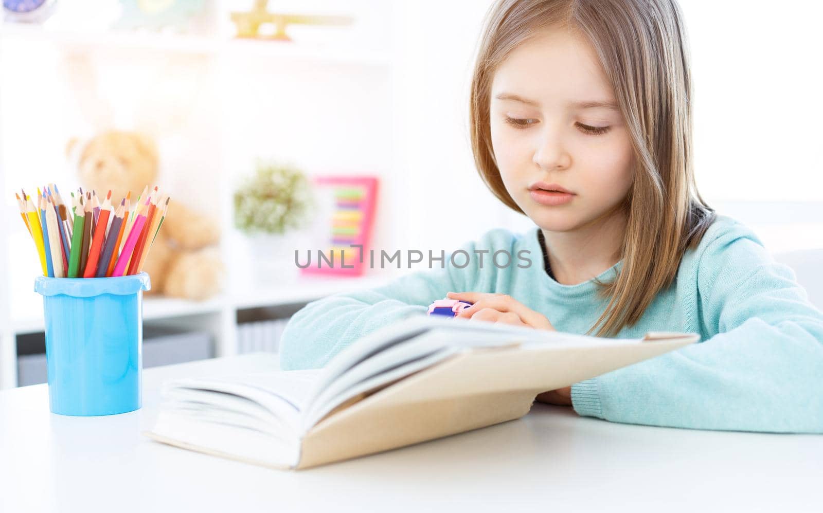 Cute little girl reading book on desk in bright room