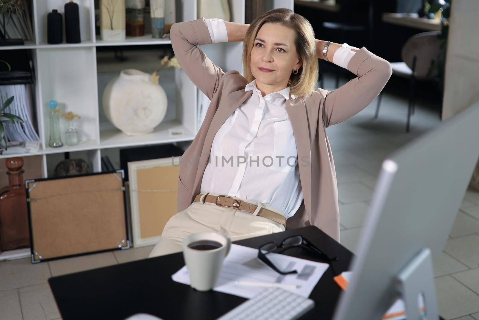 Woman with satisfied expression at desk. Arms up and folded behind her head in modern office.