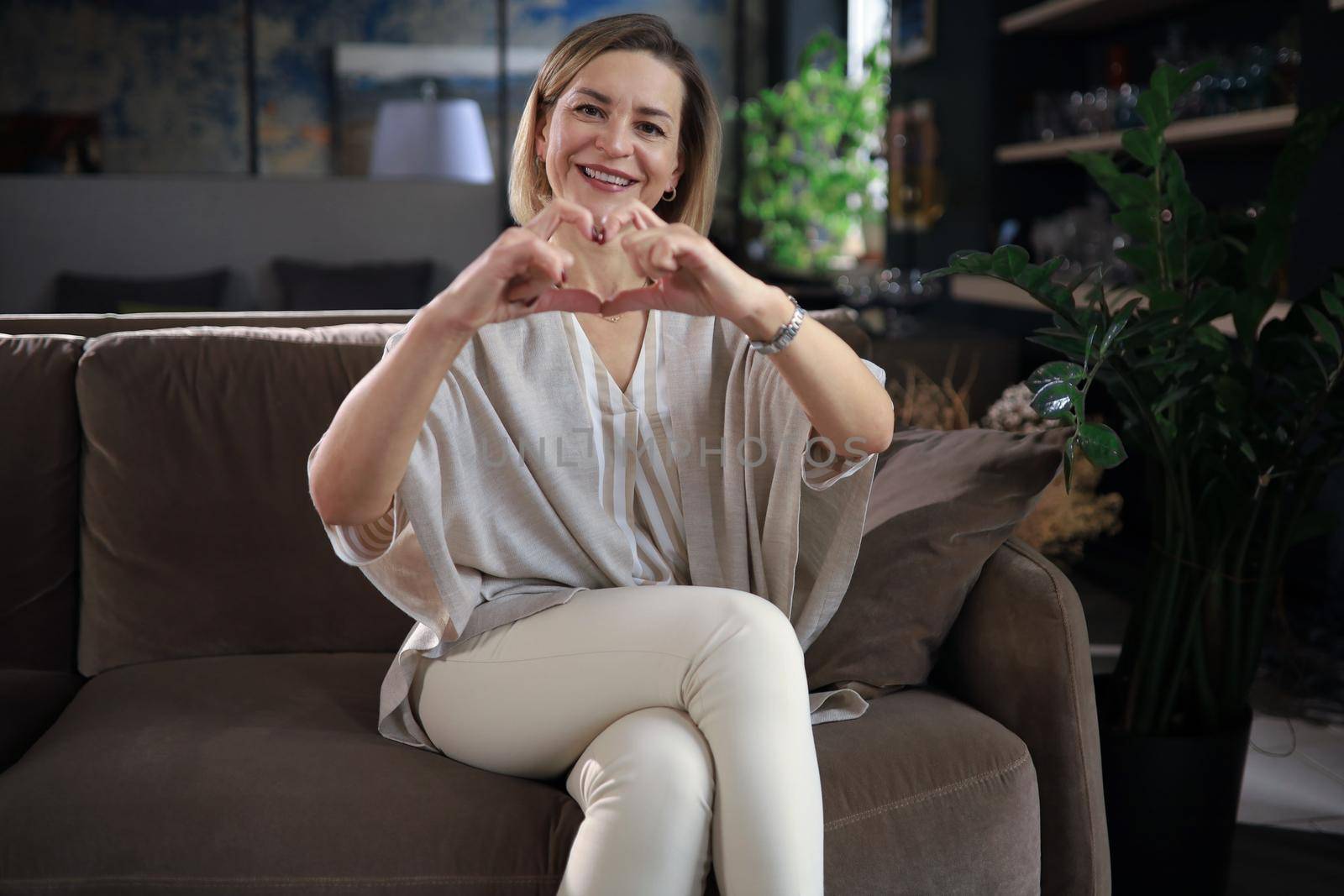 Smiling middle aged woman is showing heart gesture with two hands and looking at camera sitting on sofa