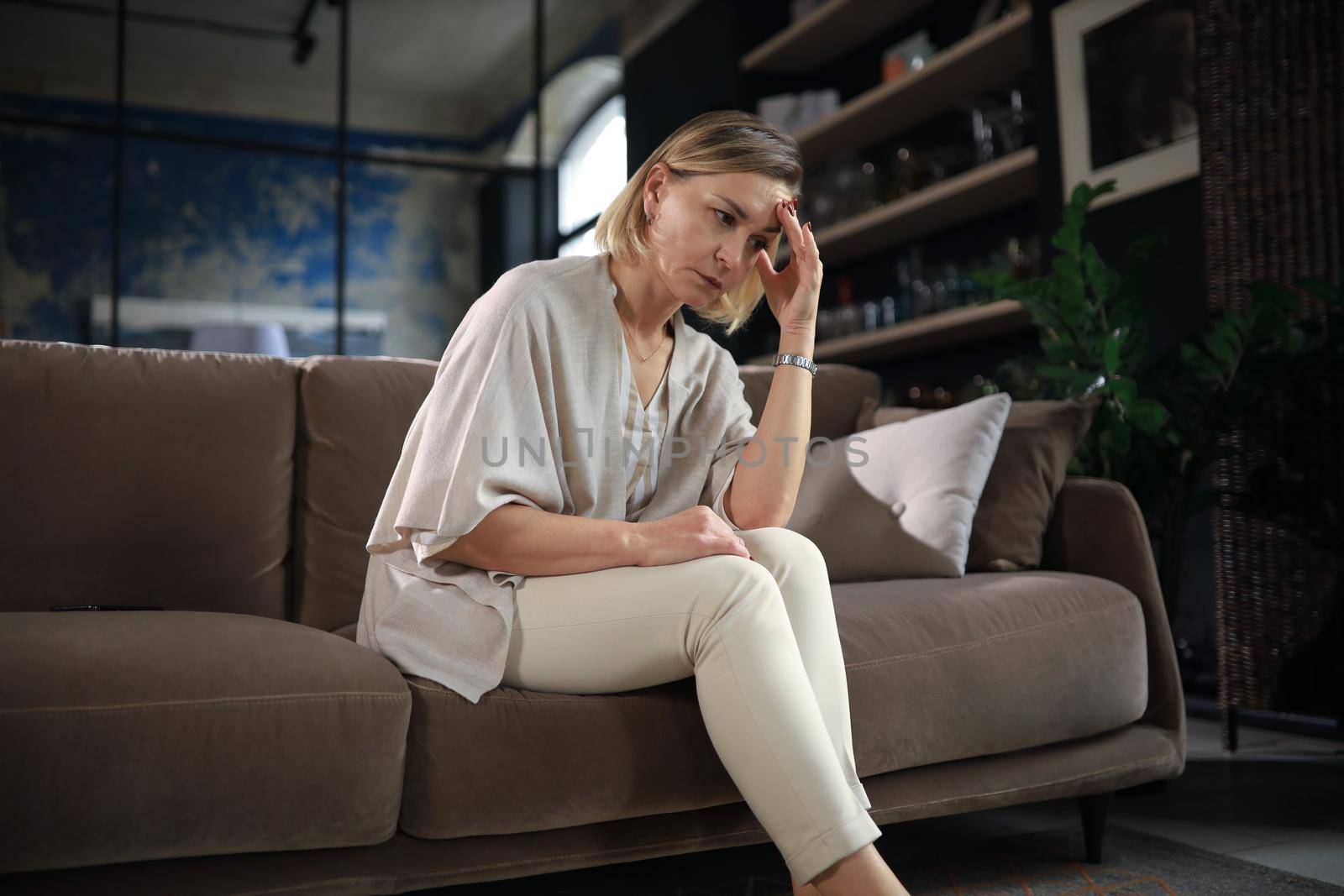 Stressed middle aged woman sit on sofa in living room, lost in thought.