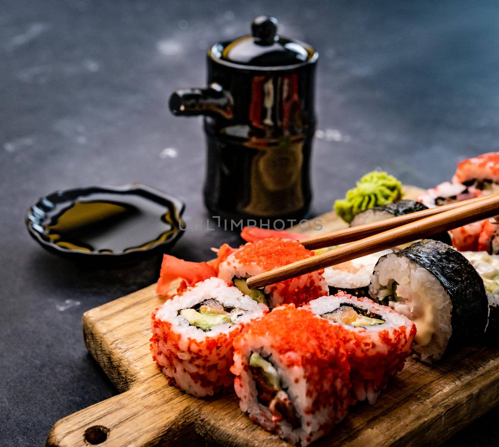 Closeup view of sushi maki set served on wooden tablet with soy sauce in bowl and person hand holding chopstick. Japanese asian roll food.