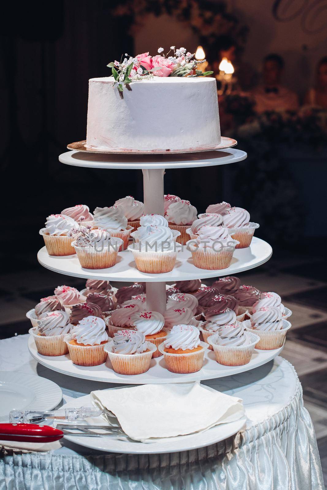 Front view of beautiful wedding cake with flowers staying on table. Delicious dessert for celebration. Lovely pair at background. Concept of love, confectionery and biscuit.