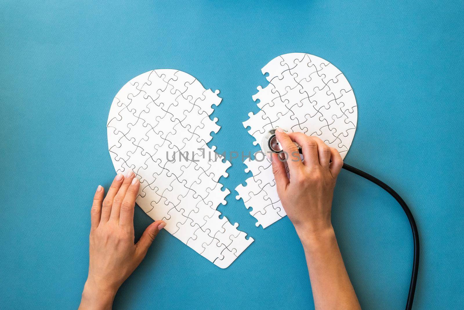 Hands with stethoscope and white puzzle in heart shape. White details of puzzle on blue background. Heart health concept.