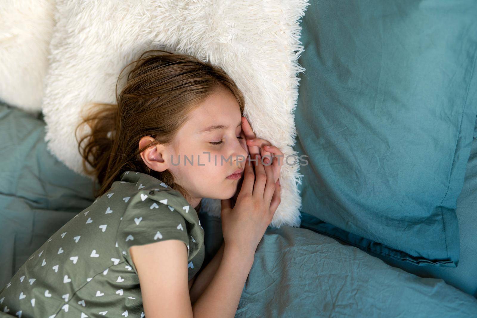 Little beautiful kid girl with blond hair lying in the bed and sleeping in bedroom on white furry pillows. Female child resting napping at home