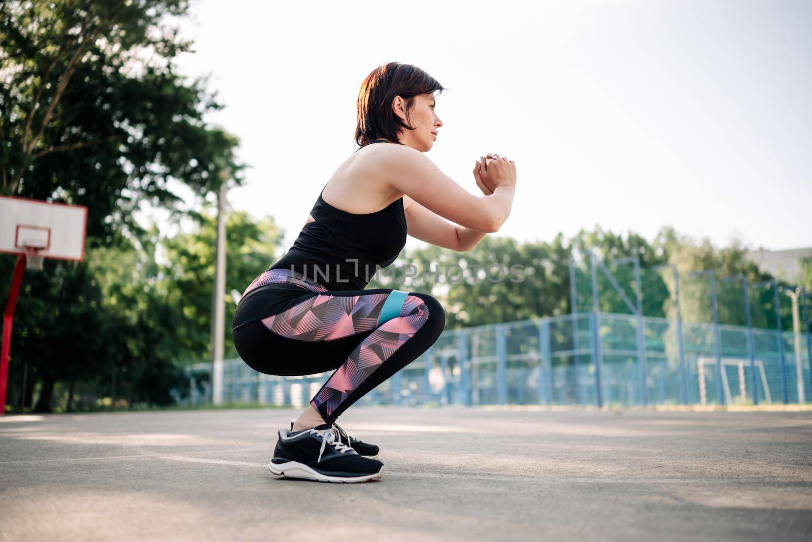 Young girl woman exercising outdoors with mini rubber elastic band doing squats at stadium. Active training for female legs and buttocks with additional sport equipment outside
