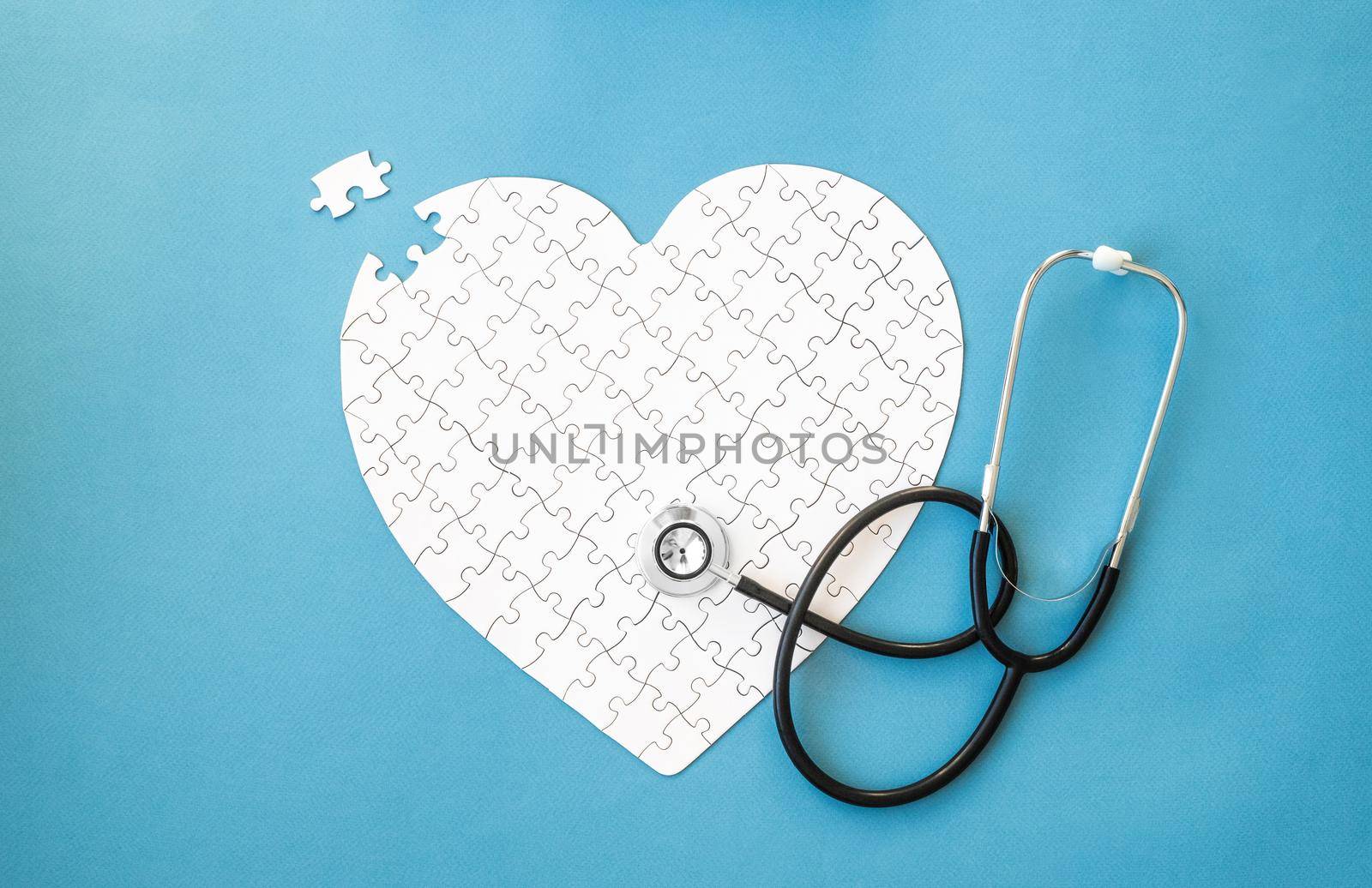 White puzzle in heart shape and stethoscope on blue background. Heart health concept