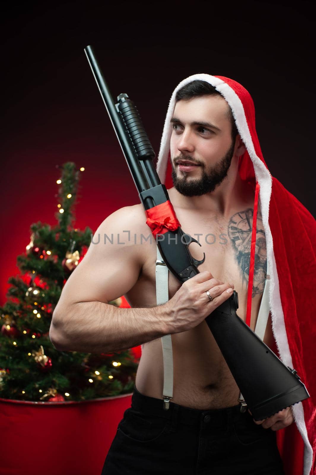 a strong guy in a red Santa Claus cape at the Christmas tree with a shotgun in his hands by Rotozey