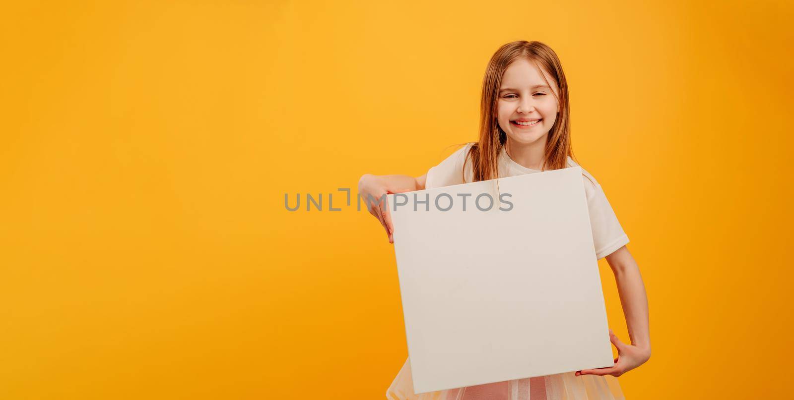 Beautiful girl child holding white canvas and looking at the camera isolated on yellow background with copyspace. Horizontal portrait of kid holding linen with place for text and smiling