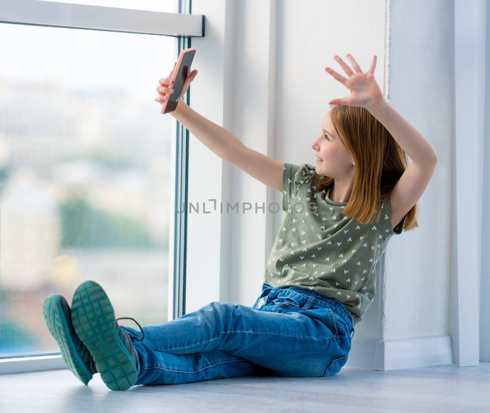 Cute preteen girl sitting on windowsill with smartphone and making selfie using stick. Beautiful female kid schoolgirl posing with cell phone at home
