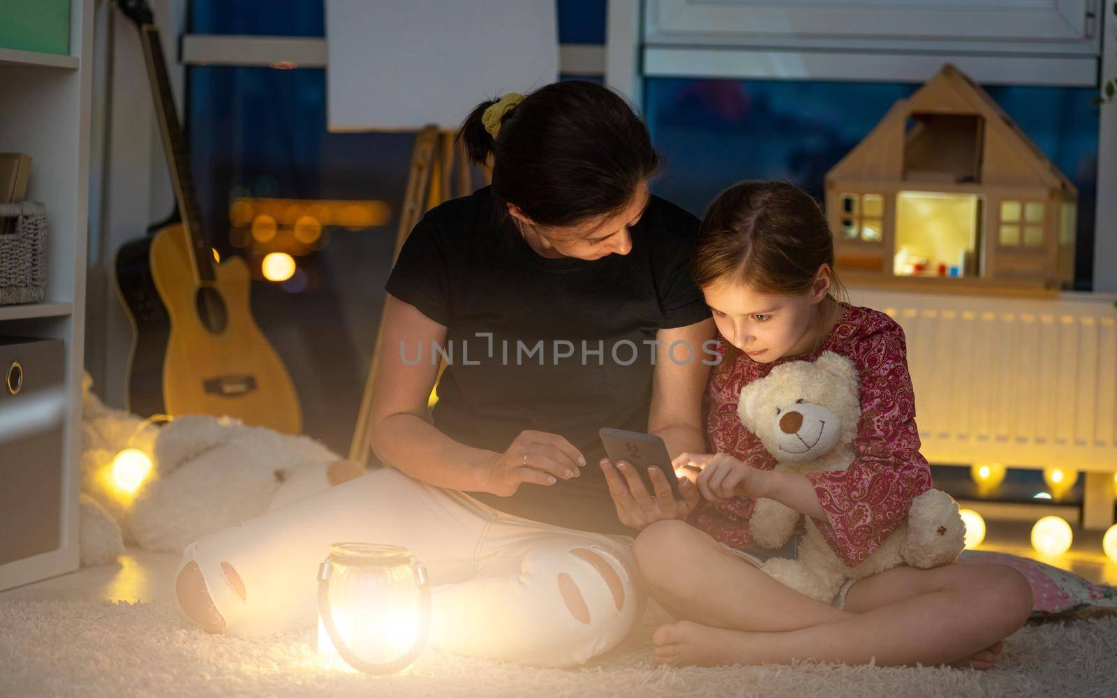 Mother showing phone to little daughter sitting on floor in playroom at night