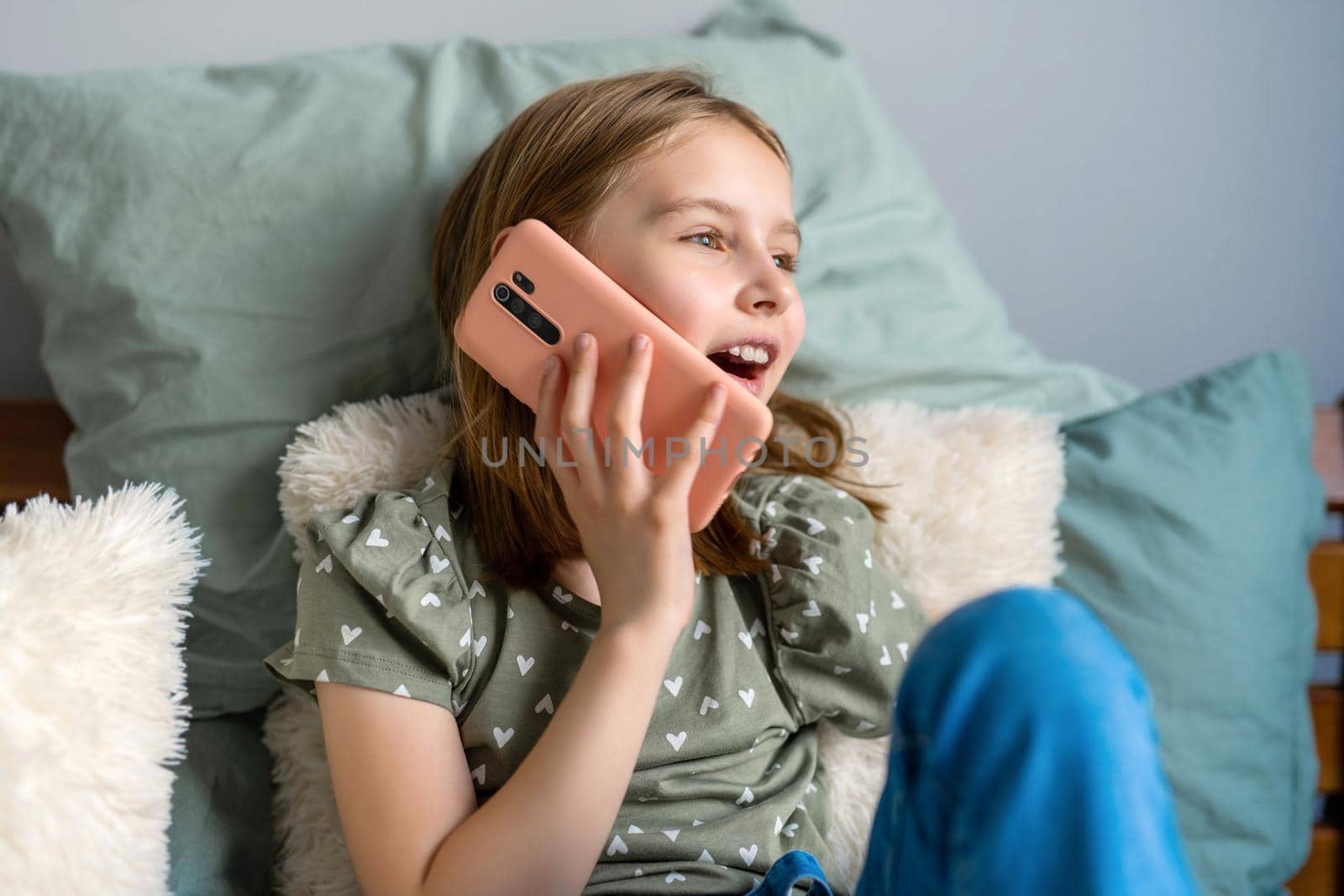Pretty preteen girl sitting in the bed and talking by smartphone at home in the bedroom. Beautiful kid communicating indoors using modern cell phone gadget